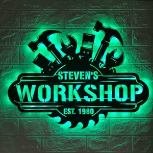 May include: A black metal wall sign with bright green LED backlighting. The design features silhouettes of a hammer, wrench, and other tools above a central banner with the text STEVEN'S WORKSHOP and EST. 1980, framed by a circular saw blade.