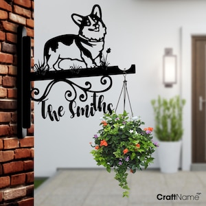 May include: Black metal sign with a silhouette of a corgi dog and the text "The Smiths". The sign is mounted on a brick wall and has a hanging planter with flowers.