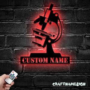 May include: A black metal wall sign shaped like a microscope with a red LED backlight. The sign features a rectangular base with the text CUSTOM NAME cut out in bold capital letters. A small remote control is shown to adjust the lighting settings.
