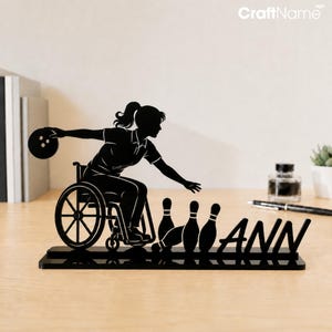 May include: A black silhouette tabletop sign featuring a person in a wheelchair bowling a ball toward three pins. The design includes the name ANN in bold capital letters integrated into the base. The entire piece is crafted from a flat material.