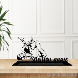 Op de afbeelding: Zwarte metalen silhouet van twee mensen in een grappling-positie, met de tekst "custom name" eronder. Het silhouet staat op een zwarte basis, die op een houten oppervlak staat.