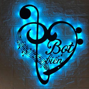 May include: A black metal wall sign shaped like a heart combined with a treble clef and musical staff lines. The design features the text "Bọt biển" in a cursive script. The sign is illuminated from behind by a bright blue LED light.