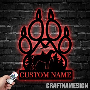 May include: A black metal paw print with a dog silhouette, trees, and a bird inside. The paw print has red LED lights around the edges. The text "CUSTOM NAME" is below the paw print. The text "CRAFTNAMESIGN" is below the name.