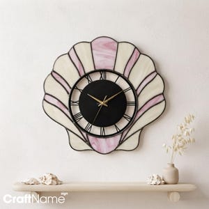 May include: A wall clock shaped like a seashell, featuring stained glass panels in cream and soft pink hues. The center is a circular black face with black Roman numerals and thin gold-colored clock hands, all framed by dark metal soldering.