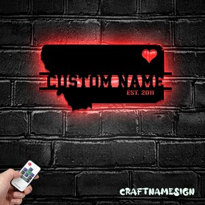 May include: A black metal wall sign shaped like the state of Montana with a red LED backlight. The sign features a cutout heart symbol, the text CUSTOM NAME, and the text EST. 2011. A small remote control is visible in the lower left corner.