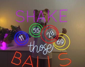 May include: A neon sign featuring the text SHAKE in purple, those in white, and BALLS in orange. Interspersed are neon circles in green, red, blue, and yellow containing the numbers 55, 28, 11, and 69 respectively, mounted on a clear acrylic backing.