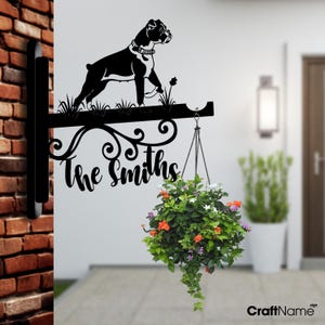 May include: Black metal house sign featuring a silhouette of a boxer dog and the text "The Smiths". A hanging planter with colourful flowers is attached to the sign.