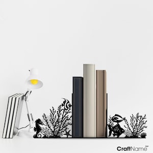May include: A black metal bookend set featuring intricate cutouts of seahorses, tropical fish, coral reefs, and starfish. The design supports a row of books, with the text CraftName sign visible in the bottom right corner of the image.