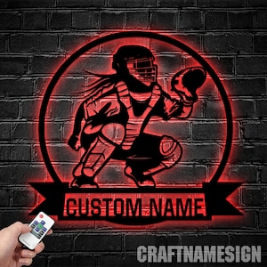 May include: A circular black metal wall sign featuring a silhouette of a baseball catcher in a crouched position, illuminated by a red LED light. A banner at the bottom contains the text CUSTOM NAME in bold, capital, sans-serif lettering.