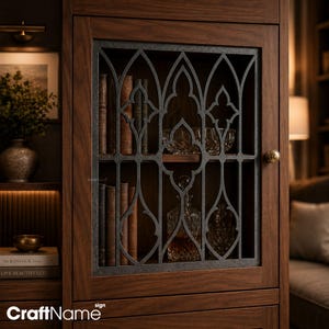 Puede incluir: Un armario de madera oscura con una puerta de cristal y un revestimiento de metal gris oscuro con un patrón gótico de arco apuntado. En el interior, los estantes contienen varios libros antiguos, un cuenco de cristal y una jarra de vidrio.