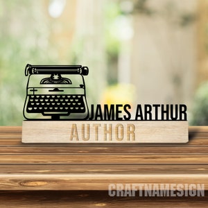 May include: A wooden desk sign with a black typewriter silhouette and the name "JAMES ARTHUR AUTHOR" cut out. The sign is made of light-coloured wood, designed for an author's desk. The background is blurred, suggesting an office or home setting.