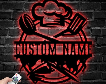 May include: A black metal wall sign featuring a chef hat, a spoon, and a spatula crossed over a circular frame with the text CUSTOM NAME in the centre. The sign is illuminated by a red LED light glow and is controlled by a small remote device.
