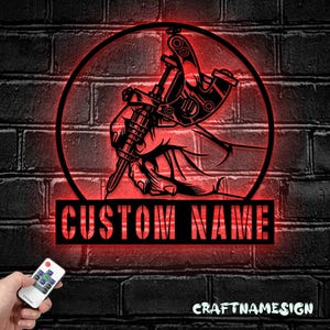 Könnte beinhalten: Ein kreisförmiges Wand-Schild aus schwarzem Metall mit einer grafischen Darstellung einer Hand, die eine Tätowiermaschine hält, beleuchtet durch rotes LED-Licht. Unter dem Kreis zeigt ein schwarzes Rechteck den Text CUSTOM NAME in Rot.