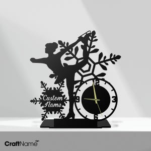 May include: Black metal clock featuring a figure skater in a graceful pose, integrated with a clock face and snowflake design. The clock includes the text "Custom Name" and is mounted on a rectangular base. The clock hands show the time.