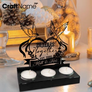 Puede incluir: Portavelas de aniversario negro cortado con l&aacute;ser con el texto "20 Years Together" y los nombres "Matt & Kate". El dise&ntilde;o presenta dos p&aacute;jaros sobre un coraz&oacute;n. Incluye tres velas de t&eacute;.