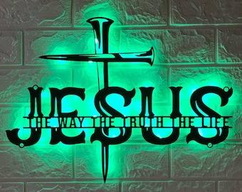 May include: A black metal sign with the word "JESUS" in large letters and the phrase "The Way The Truth The Life" in smaller letters below. The sign is illuminated with green light.