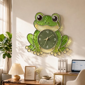 May include: A wall clock shaped like a frog, constructed from textured green and yellow stained glass with dark metal framing. The central circular clock face is dark green with white numerals at 3, 6, 9, and 12, featuring gold-colored hands.