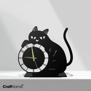 May include: A black metal desk clock shaped like a sitting cat with its paws resting on a circular clock face. The clock features a white border with black numerals and thin gold-colored hands. The cat has a heart-shaped nose and a long tail.