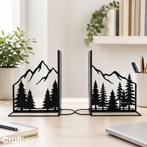 May include: A pair of black metal bookends featuring a laser-cut silhouette of a mountain range and a forest of pine trees. The design includes a flat base for stability and a vertical panel displaying the detailed landscape scenery.