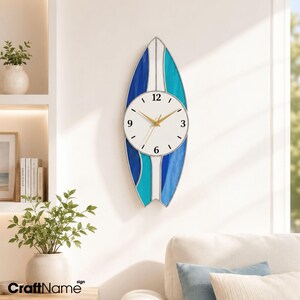 May include: A surfboard-shaped wall clock featuring a white center panel with black numerals 3, 6, 9, and 12. The clock face is surrounded by stained glass panels in shades of deep blue, light blue, and white, with gold-colored clock hands.