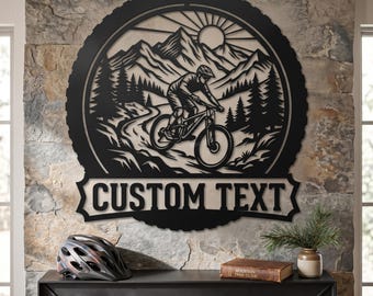 May include: Black metal wall art featuring a mountain biker on a trail, mountains, and a sun. The design is circular with a banner that reads "CUSTOM TEXT". A helmet, book, and small plant are on the mantel.