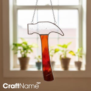May include: A stained glass hammer hangs from a chain. The hammerhead is clear, while the handle transitions from orange to red. The background shows a window with potted plants. The text "CraftName sign" is at the bottom.