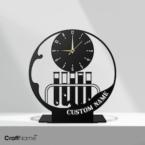May include: Black metal desk clock with a circular design. The clock face features black numbers and gold hands. Below the clock is a test tube rack. The base reads "CUSTOM NAME". The clock is a unique gift.