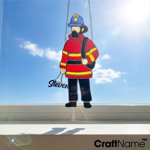 Puede incluir: Una figura de bombero de vidriera, colgada de cadenas. La figura lleva una chaqueta roja con rayas amarillas, pantalones negros y un casco azul. El nombre "Steven" está escrito en la figura. El letrero dice "CraftName sign".