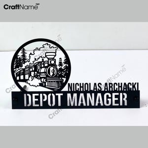 May include: Black metal sign with a train and trees in a circle. The sign reads "Depot Manager" and "Nicholas Archacki".