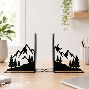 May include: A pair of black metal bookends featuring a laser-cut silhouette design. One piece depicts a mountain range with pine trees, while the other shows a mountain range with pine trees and a crescent moon. The text CraftName is visible.