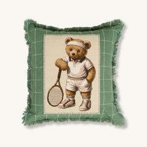 Op de afbeelding: Een decoratief vierkant kussen met een tennisthema. Het heeft een kruissteekontwerp van een bruine teddybeer in tennisoutfit met een racket. Het kussen heeft een groene geruite rand met een gefranjerde rand.