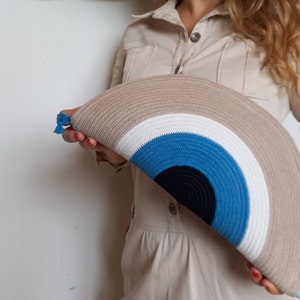 May include: A large, round, crocheted clutch bag with a rainbow pattern in shades of beige, white, blue, and black. The bag has a blue tassel.