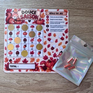 Peut inclure: Un tableau de défi d'épargne rectangulaire intitulé Douce Saison avec une grille illustrée de citrouilles, champignons et glands, accompagné d'une pochette holographique contenant quatre petites gélules rouges et blanches pour un jeu d'épargne.