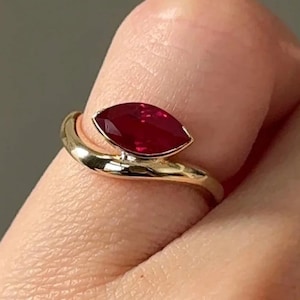 May include: A gold ring featuring a marquise-cut, deep red gemstone. The ring's band curves around the finger, creating a modern and elegant design. The gemstone is set in a bezel setting, enhancing its vibrant color and shape.