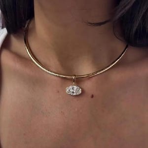 May include: A gold-colored necklace with a pendant. The pendant is an oval shape with a cluster of clear stones. The necklace is a choker style, sitting close to the neck.