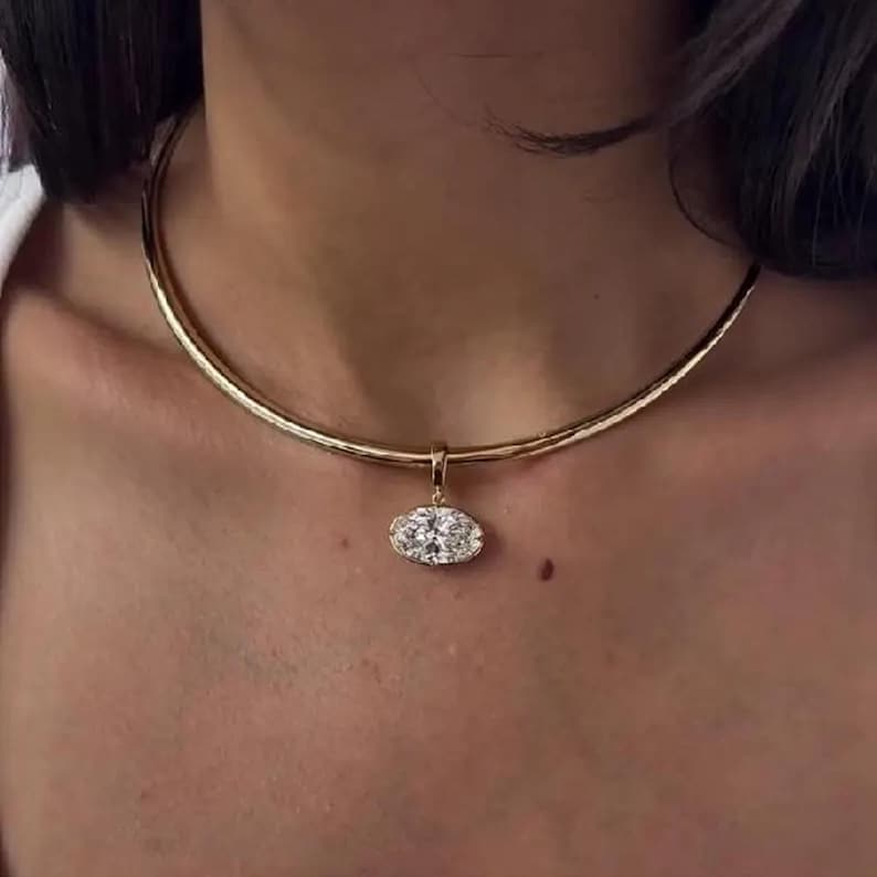 May include: A gold-colored necklace with a pendant. The pendant is an oval shape with a cluster of clear stones. The necklace is a choker style, sitting close to the neck.