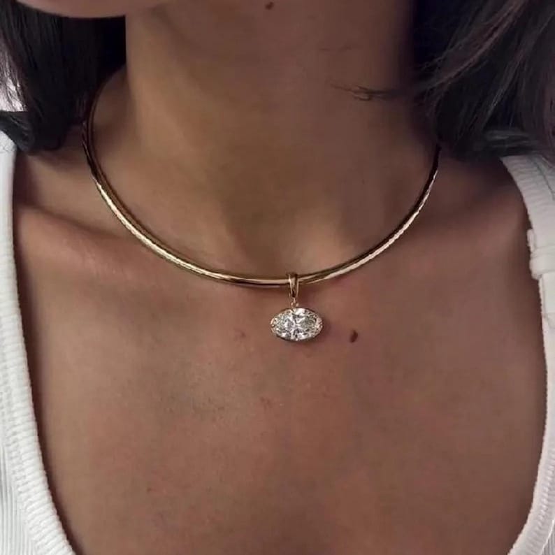May include: A gold-toned choker necklace with a pendant. The pendant is an oval-shaped, clear gemstone with a halo setting. The necklace is worn around the neck.