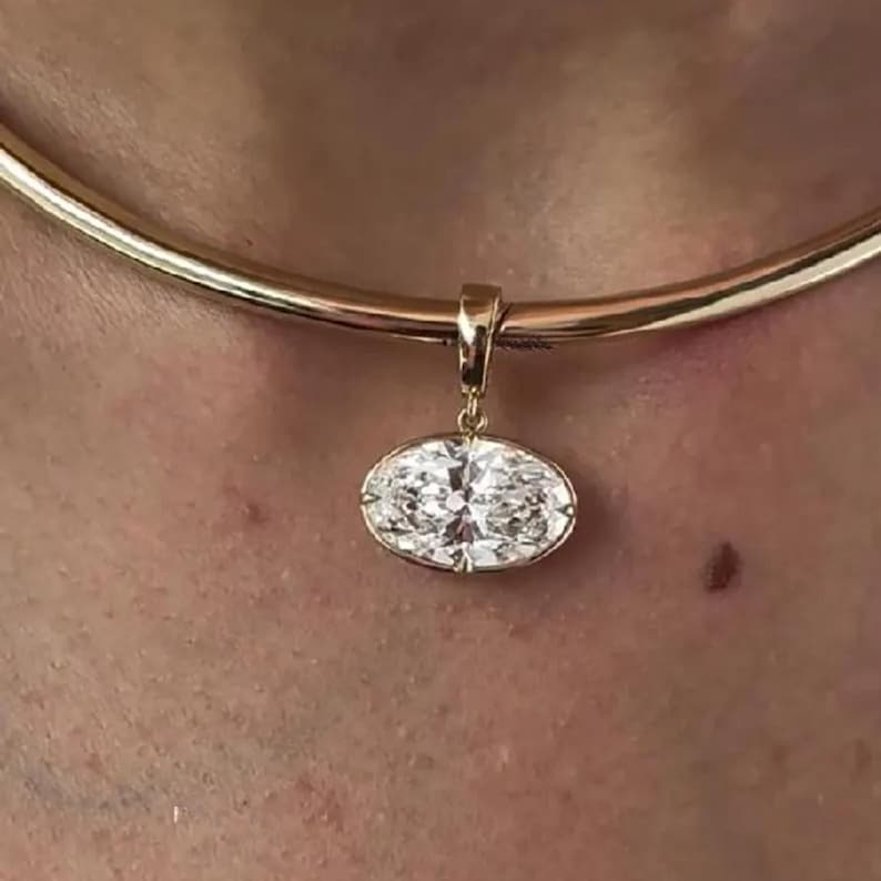 May include: A gold-colored necklace with a large, oval-shaped diamond pendant. The diamond is set in a gold bezel and sparkles brightly. The necklace is a simple, elegant design.
