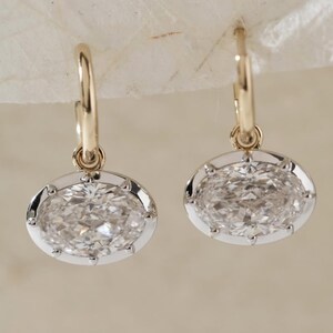 May include: A pair of gold hoop earrings with oval-shaped, clear gemstones. The gemstones are set in a silver-colored bezel with small decorative prongs. The earrings are displayed against a neutral background.