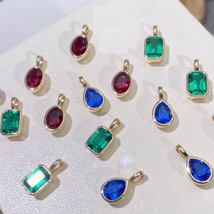 May include: A collection of gold-toned pendants with various gemstone shapes and colors. The pendants feature emerald green, ruby red, and sapphire blue stones. The gemstones are set in gold bezels, showcasing their vibrant hues.