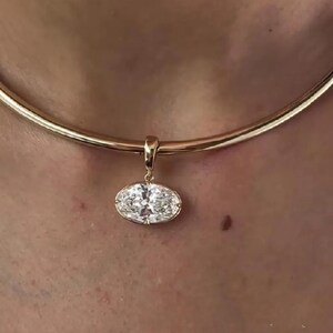 May include: A gold-toned necklace with a pendant featuring a large, oval-cut diamond. The pendant hangs from a simple, curved gold chain. The diamond sparkles with multiple facets, reflecting light.