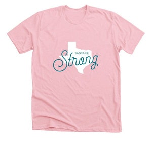May include: Pink t-shirt with a white outline of the state of Texas and the text "Santa Fe Strong" in teal blue.