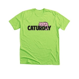 Puede incluir: Una camiseta verde lima con una ilustración de un gato rosa y el texto "CATURDAY" en negro.