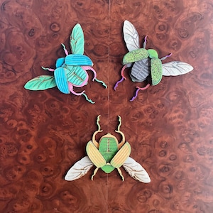 May include: Three colorful wooden beetle ornaments with intricate details. The beetles are green, blue, and pink with black accents.