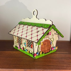 May include: A wooden Easter house ornament with a white and pink roof, green vines, and a brown door. The house is decorated with colourful Easter eggs.