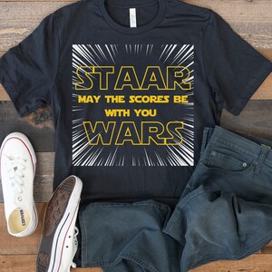 May include: A black t-shirt with a white graphic that says "STAAR May the scores be with you WARS" in a yellow font. The graphic is surrounded by white lines radiating outward.