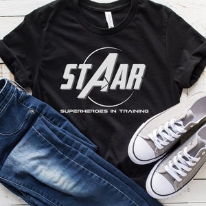 May include: Black t-shirt with a white graphic of a circle with the word "STAAR" inside and the text "SUPERHEROES IN TRAINING" below.