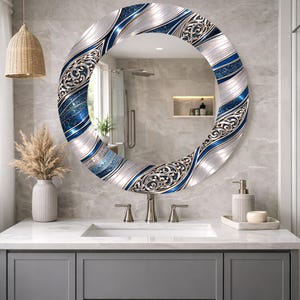 May include: An oval mirror with a decorative border. The border features silver, blue, and white swirling patterns with ornate detailing. The mirror is mounted above a bathroom sink.