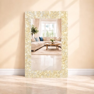 May include: A rectangular mirror with a decorative frame. The frame is made of small, irregularly shaped, off-white and gold-toned pieces. The mirror reflects a living room with a beige sectional sofa, a wooden coffee table, and a window.