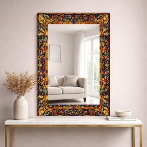 May include: Rectangular mirror with a stained glass style frame. The frame features a colorful floral and geometric design in shades of red, yellow, blue, and brown. The mirror reflects a living room with a white sofa and window.